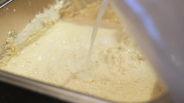High-angle close-up of fresh liquid milk being poured into a large metal bowl containing a wheat flour and yeast mixture. A professional culinary shot capturing the initial stage of making artisanal b