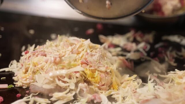High-angle shot of a chef or diner preparing Japanese okonomiyaki, a savory pancake made from wheat-flour batter, shredded cabbage, and various toppings, sizzling on a professional flat teppan griddle