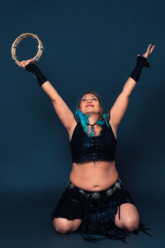 Happy woman dancer is kneeling and raising her arms up, holding tambourine in one of them. Expression woman is attired in black handmade costume, which consists of leather top and skirt, bracers