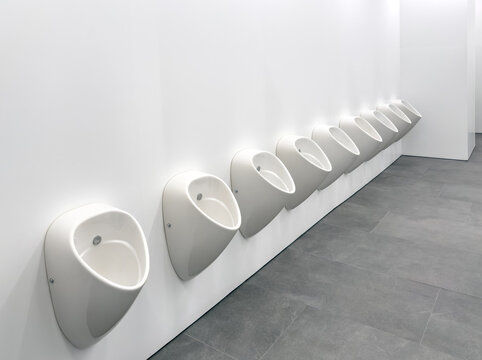Row of modern white ceramic urinals in a clean public restroom interior. Contemporary minimalist bathroom design with grey stone tiles and bright lighting in commercial space