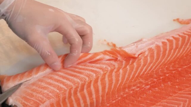 A close-up, high-angle shot of a chef's hands skillfully using a sharp knife to remove the fatty belly lining from a vibrant orange raw salmon fillet. The process demonstrates professional fishmonger 