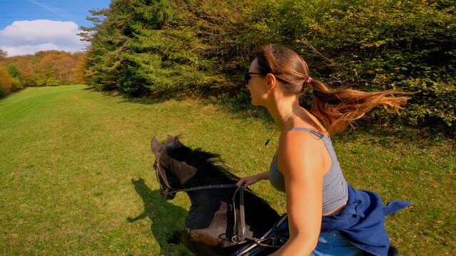 SELFIE: Woman horseback riding across a sunny meadow by the lush green forest on sunny day. Smiling female rider galloping with her dark brown stallion through the beautiful countryside in springtime.