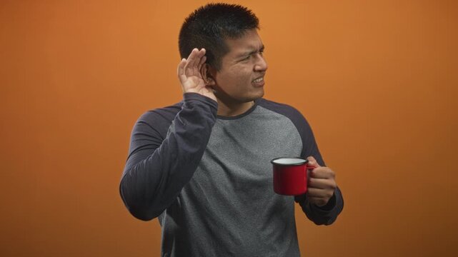 Man holding red enamel mug, cupping his ear with hand and squinting while standing against an orange studio wall; curiosity listening.