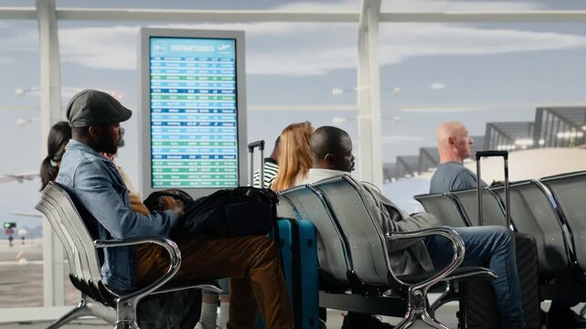 Travelers walking in busy airport to boarding gate after seeing departure announcement on FIDS display. Diverse tourists hearing boarding call, leaving chairs, navigating transit hub