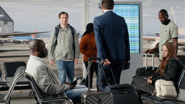 African american woman in busy airport sitting down on chair, waiting for plane. Tourists carrying trolley in terminal reading boarding gate number on departure board display, sitting on bench