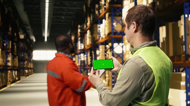 Chroma key on phone, with employee near racks, cardboard boxes in busy depot. Supply chain operations and inventory management creates import export and e-commerce industry, dispatch.