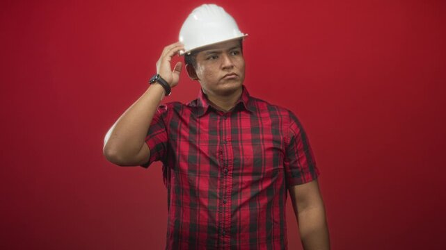 Man adjusting white hardhat with right hand to head, wearing red plaid shirt and wristwatch against studio red backdrop; thoughtful.