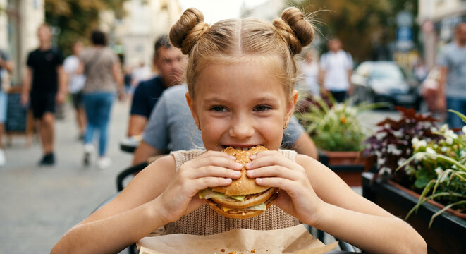 A happy girl is eating a large sandwich at a table in a street cafe