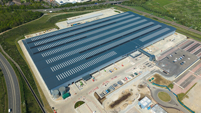 Aerial view of the large Symmetry Park distribution center with solar panels on the roof and surrounding parking areas near the A14 road in Kettering, England, United Kingdom.