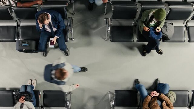 Top down view of crowd walking in busy airport, sitting on chairs. Passengers navigating chaotic transit hub environment before departure during flight layover, waiting on benches, high angle shot