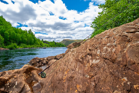 A large salmon river with multiple fishing pools and a sharp bend in the river. Moving water or rapids creates white waves. There are trees on both sides and large rocks along the riverbank. 