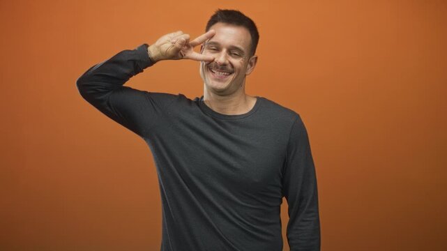Young hispanic man with moustache flashing a peace sign over his eye while smiling in a studio with an orange backdrop; playful joy.