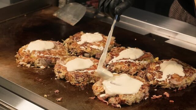 Close-up video of fresh Japanese okonomiyaki savory pancakes being prepared on a professional teppanyaki iron plate. The chef is drizzling creamy mayonnaise over the golden-brown crust. A popular stre