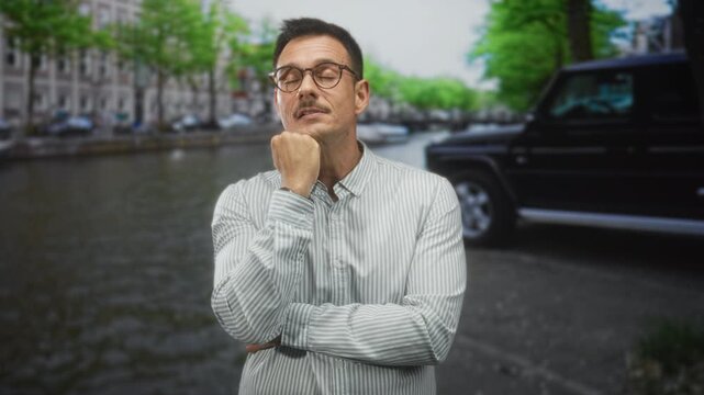 Hispanic man with moustache and glasses yawning with hand on chin beside a canal street with parked suv and trees; fatigue contemplation.
