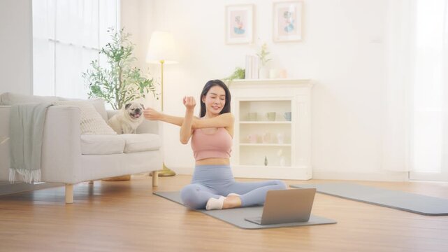 Asian woman stretching body during online yoga class workout exercise on yoga mat using laptop at home, healthy lifestyle fitness routine and indoor wellness body care with pug dog pet in living room