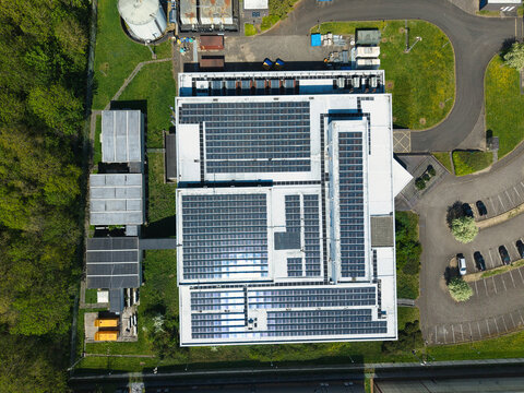 Aerial view of the solar panel arrays on the roof of a modern data centre surrounded by lush green trees and industrial infrastructure in Wellingborough, England, United Kingdom.