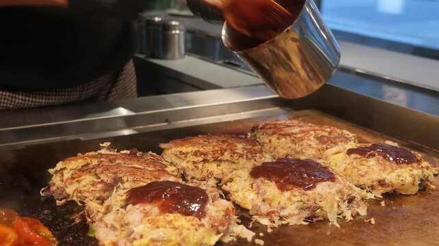 A close-up shot of a chef's hand pouring a thick, savory brown sauce from a metal container onto several okonomiyaki pancakes cooking on a hot iron teppan grill in a traditional Japanese restaurant se