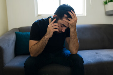 Obraz premium Stressed man sitting alone on sofa making a phone call, feeling lonely and seeking support for depression
