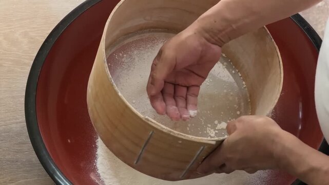Close-up footage of a person traditionally sifting fine buckwheat flour through a circular wooden sieve into a large, red-and-black lacquer konebachi kneading bowl. This authentic Japanese culinary pr