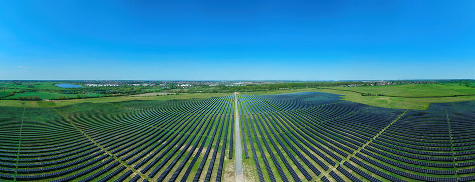 Aerial view of a large solar farm with rows of photovoltaic panels stretching across green fields under a clear blue sky in Little Irchester, England, United Kingdom.