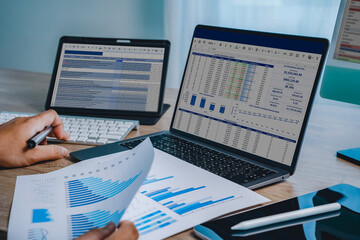 Hands reviewing financial chart document with laptop displaying spreadsheet dashboard and tablet screen, representing data analysis workflow, reporting process, business performance tracking, and digi