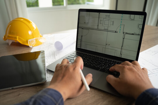 Hands using laptop displaying architectural blueprint with stylus and devices on desk, representing AI assisted design workflow, engineering data analysis process, digital modeling, and construction p