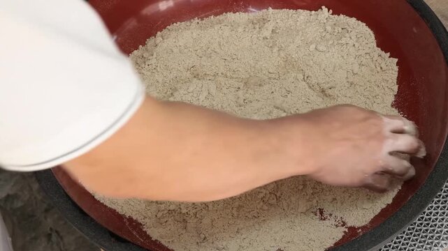 close-up shot of a person&rsquo;s hand pouring water into a large red bowl filled with coarse whole wheat flour (atta). The video captures the initial step of the traditional dough-kneading process for maki