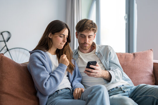 A young couple sits close on a living room sofa, focused on a smartphone as they read messages. The pair appear concerned and engaged, sharing a quiet, emotional moment at home.
