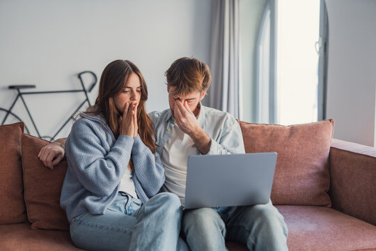 Young couple sits on a sofa looking shocked and worried while viewing something upsetting on a laptop, conveying stress, anxiety, relationship tension and financial or personal bad news at home.