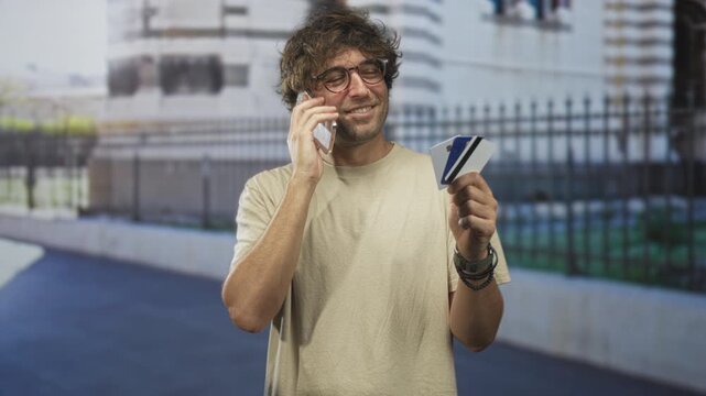 Man holding creditcard and phone to ear, smiling while talking and displaying cards near a stone building and iron fence on a street; confidence convenience.