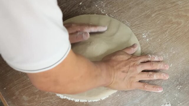 High-angle close-up of a professional chef's hands skillfully pressing and stretching a round piece of fresh, floured pizza dough on a rustic wooden countertop. The footage captures the traditional "s
