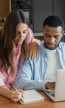 A couple collaborates at a kitchen table using a laptop and notebook to plan finances and projects, sharing ideas and notes in a warm home office setting focused on teamwork and organization.