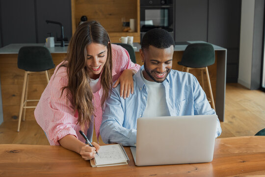A happy couple collaborates at their kitchen table, using a laptop and notebook to plan finances, manage bills and work from home while sharing ideas in a relaxed modern home environment.