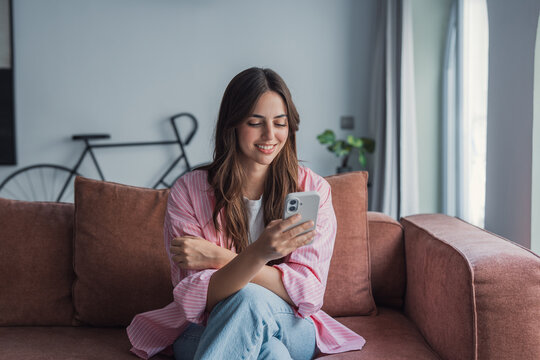 Young woman sits comfortably on a sofa in a bright living room, smiling as she uses her smartphone. Casual, relaxed moment conveying connection, leisure, home life and modern communication.