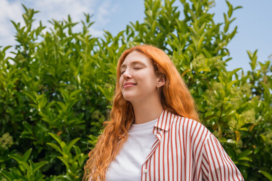 A young redhead woman stands outdoors with eyes closed, breathing deeply and smiling softly amid lush green foliage, conveying calm, contentment and a relaxed summer vibe in casual striped shirt.