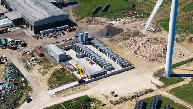 Aerial view of Chelveston Wind Farm featuring battery storage containers, solar panels, and a large wind turbine near industrial buildings in Wellingborough, England, United Kingdom.