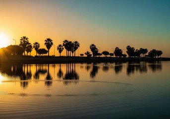 Obraz premium Tropical palm trees stand in silhouette against a vibrant golden sunset reflecting on calm water.