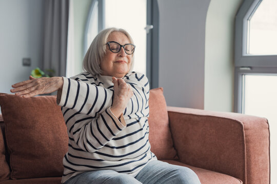 A relaxed senior woman wearing glasses stretches her arms on a comfortable sofa in a bright living room, enjoying gentle mobility exercise and calm moments of self-care and healthy aging.