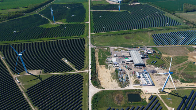 Aerial view of Chelveston Wind Farm featuring wind turbines and vast solar panel arrays across green fields in Wellingborough, England, United Kingdom.