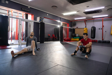 Fototapeta premium Diverse male training partners sitting on mats at MMA gym cage, wearing rashguard and gloves