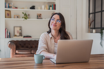 Fototapeta premium Young woman sits at a wooden desk in a cozy living-room home office, working on a laptop and smiling thoughtfully with a coffee mug nearby, conveying relaxed remote work, creativity and focus.