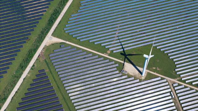 Aerial view of Chelveston Wind Farm featuring rows of solar panels and a large wind turbine casting a shadow over green fields in Wellingborough, England, United Kingdom.