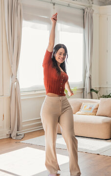 A happy young woman dances in a sunlit living room, smiling and stretching her arm up. Casual outfit and glasses convey a relaxed, confident lifestyle and joyful morning at home.