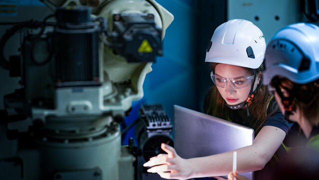 Two female engineers in hard hats and safety vests collaborate in a dark factory. They are focused, looking at a high tech machine or screen during a night shift.