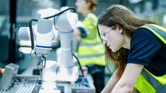 Two female engineers in safety vests collaborate in a tech lab. One woman in gloves inspects a high tech robotic arm while her colleague holds a tablet.