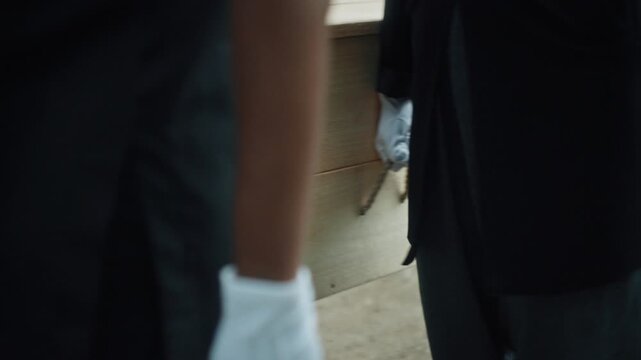 Pallbearers in black suits and white gloves carrying a simple wooden casket during a burial service