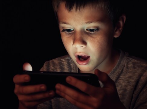 Young boy stares wide eyed at smartphone screen in dark room. Child engrossed by glowing mobile device. Youth absorbed in digital world, possibly gaming or watching videos.
