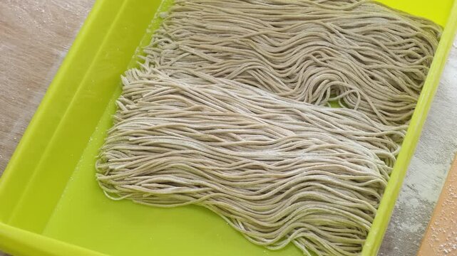 A close-up shot showing the traditional process of handling fresh, flour-dusted noodles in a yellow tray. The hands of an artisan are seen carefully organizing and lifting the strands of dough, highli