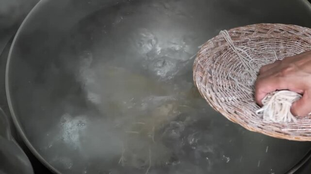 A close-up shot showing bundles of thin rice vermicelli noodles being transferred from a woven basket into a large metal pot of boiling water with visible steam rising. The process highlights traditio