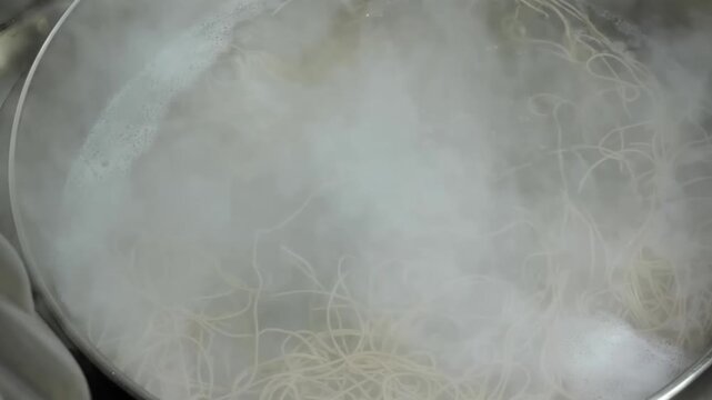close-up shot of thin noodles or spaghetti cooking in a pot of rapidly boiling water. Thick white steam and mist rise from the surface, creating an atmospheric and warm culinary scene. Ideal for cooki